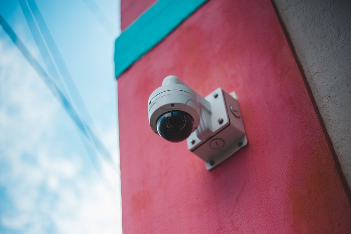 A surveillance camera mounted on a vibrant pink and teal wall outdoors.