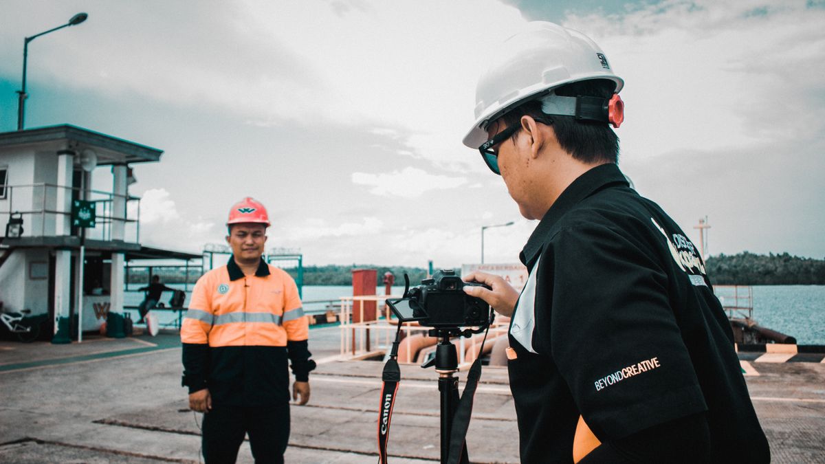 Two construction workers in safety gear filming at a waterfront location.